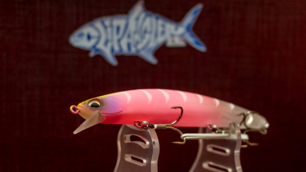 Un'esca artificiale Tide Minnow Sprat con finiture rosa e gialla, posizionata su un supporto trasparente, con un logo sullo sfondo.